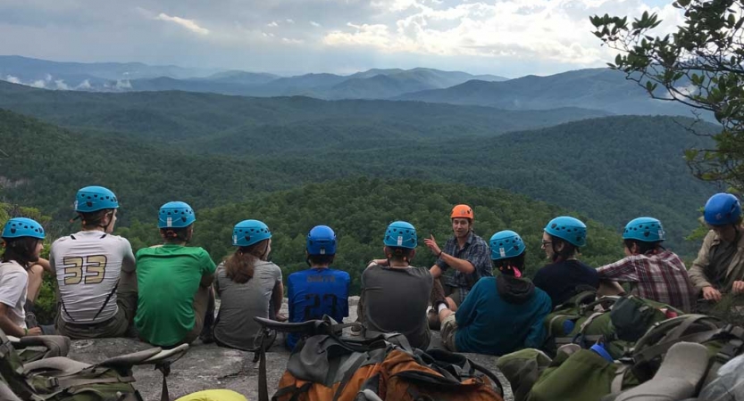 A group of students wearing helmets sit in a line and listen to an instructor speak. In the background, there is a vast and green mountainous landscape.